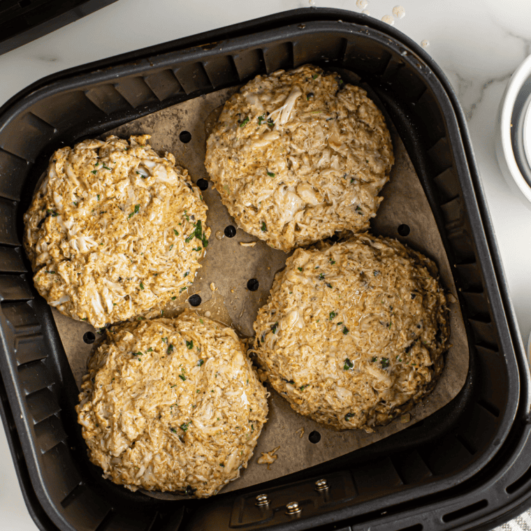 Crab cake patties cooking in an air fryer basket, golden and crisping while being lightly sprayed with oil.