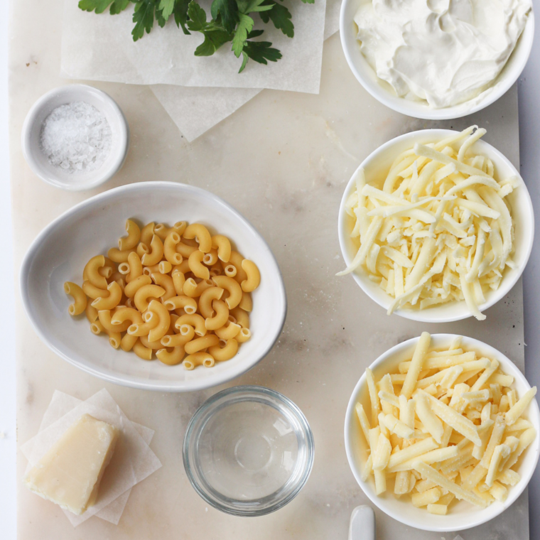 Ingredients for Panera mac and cheese arranged on a table, including elbow macaroni, shredded cheddar cheese, cream, butter, and seasonings.