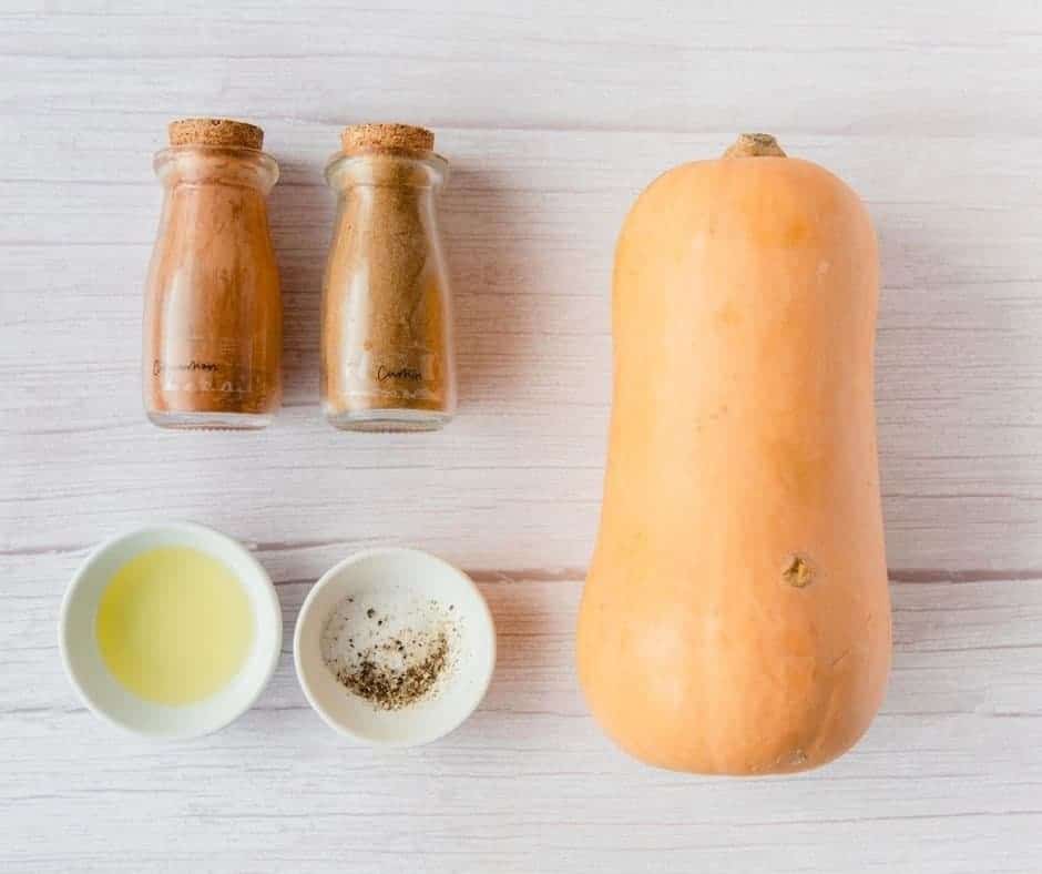 Top view of Butternut Squash, a ramekin of Olive Oil, a ramekin of Ground Cumin and Ground Cinnamon, and a shaker of Salt, and one of Black Pepper lying down on a worktop. 