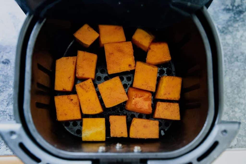 Top view of Butternut Squash being chopped into cubes on a wooden chopping board, with a knife lying next to some of the cubes. 