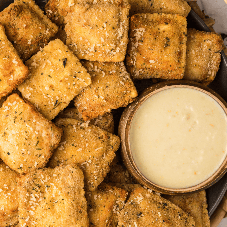 Toasted ravioli arranged on a platter with a bowl of creamy Alfredo dipping sauce