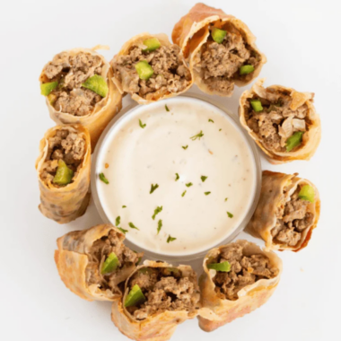 TGI Fridays Philly cheesesteak egg rolls sliced on the diagonal and stacked on a white plate with creamy horseradish dipping sauce