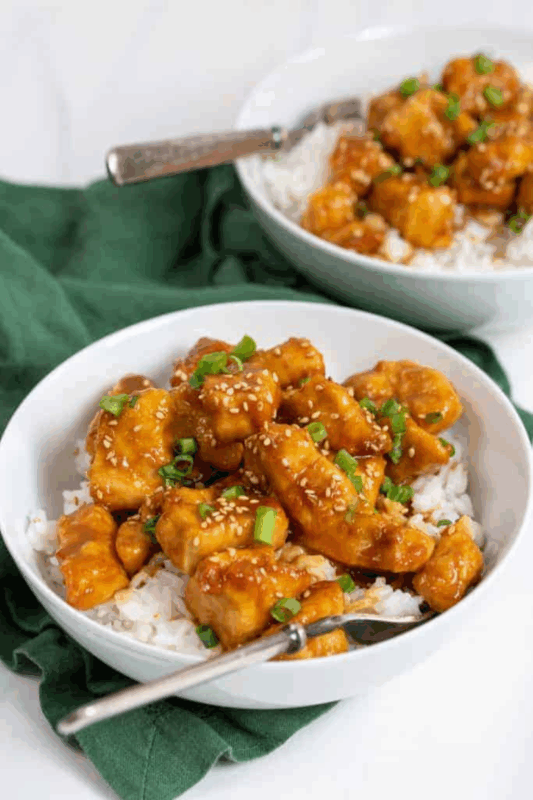 Crispy sesame chicken coated in glossy sauce, garnished with sesame seeds and green onions on a plate.