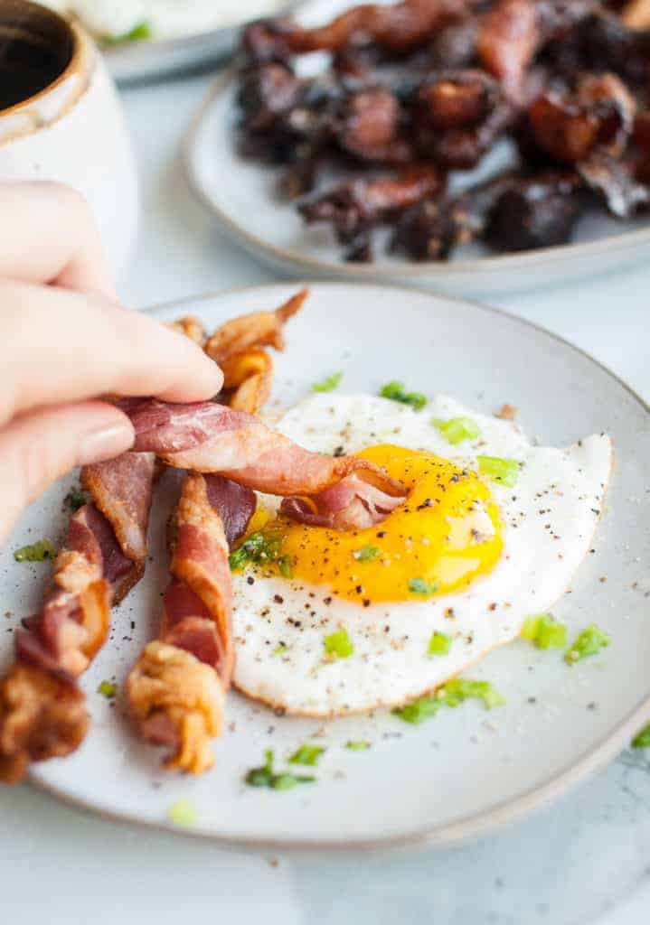 A white plate with a fried egg and two twisted strips of bacon on it. There is a hand lifting a third twisted strip of bacon and dipping it into the fried egg's yolk. 
