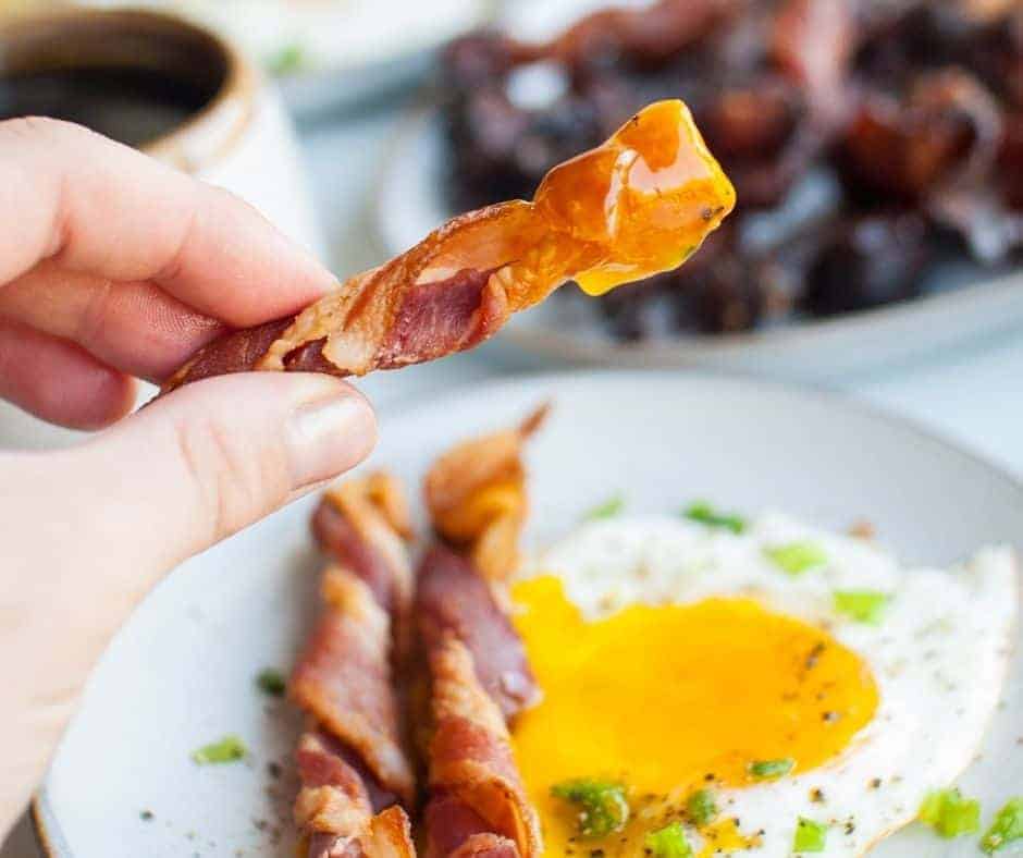 White plate in the background with fried eggs and some air fryer twisted bacon. In the foreground there is a hand lifting a piece of crispy air fryer bacon that has been dipped into the egg yolk.