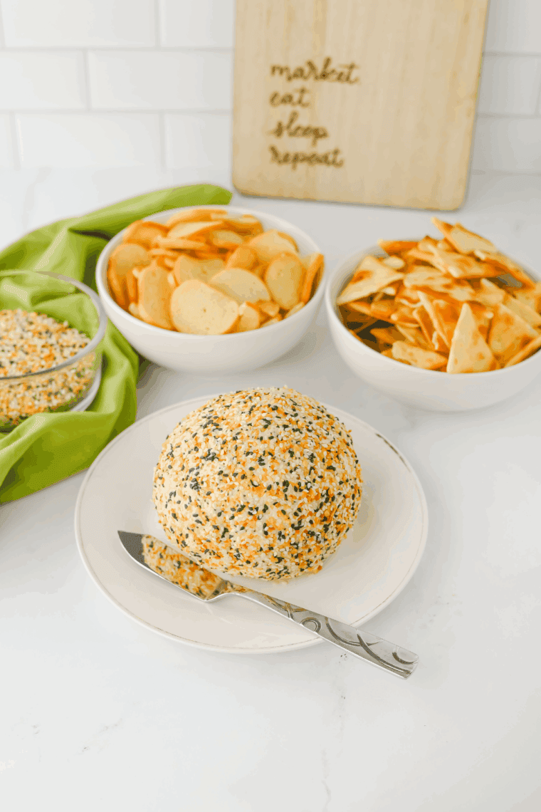 Everything Bagel Cheese Ball coated with seasoning, served with crackers and fresh vegetables for dipping.