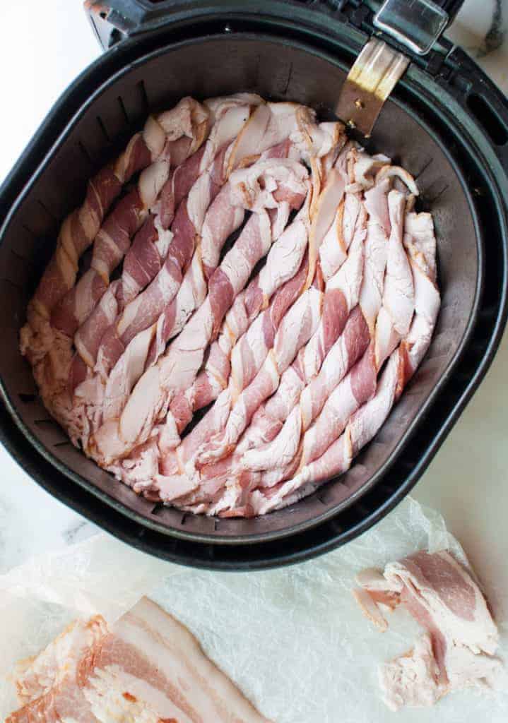 Air fryer basket filled with strips of uncooked twisted bacon. 