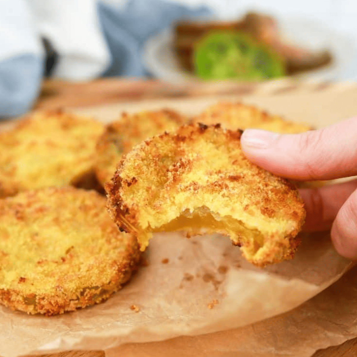 Close up of a hand holding an air fryer fried breaded green tomato, with more fried green tomatoes in the background on greaseproof paper on a chopping board. 