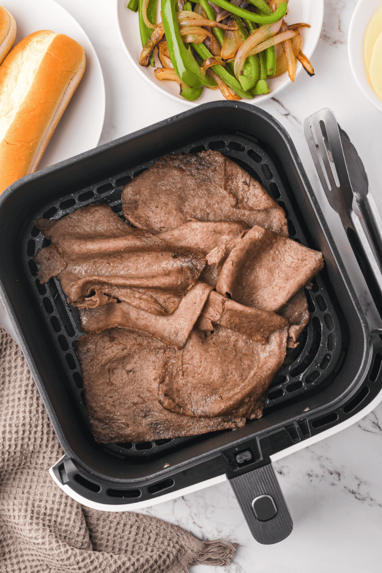 Cooking frozen Steak Umms in an air fryer basket, tossing with tongs for even cooking.