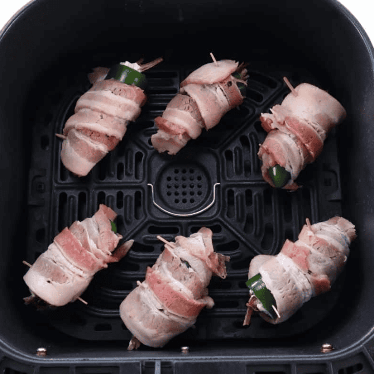 Bacon-wrapped stuffed jalapeños secured with toothpicks placed in an air fryer basket, ready to cook.
