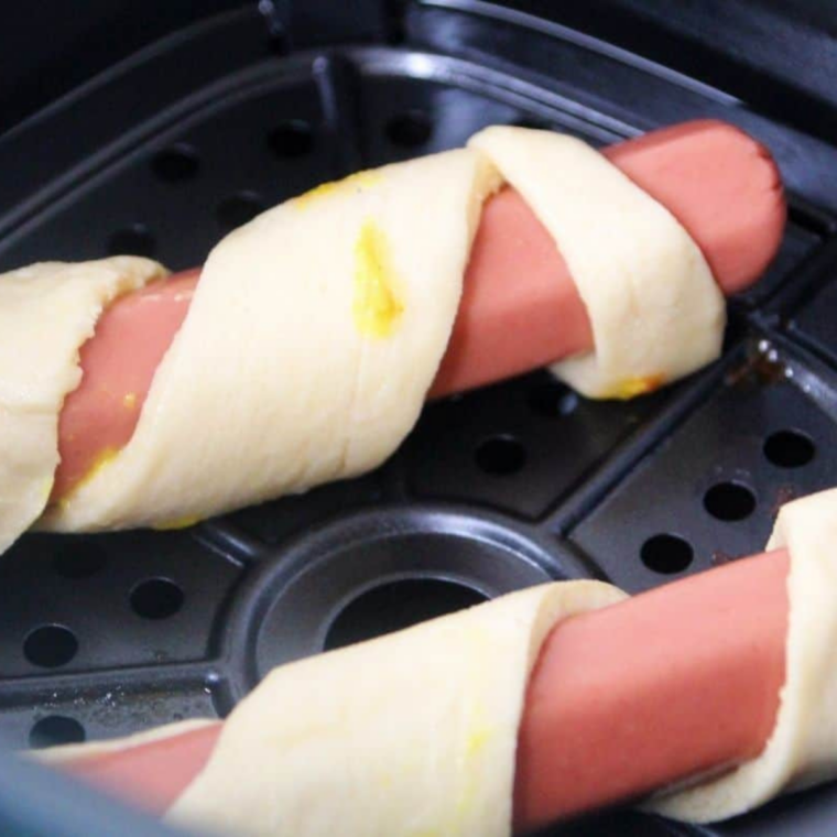 Air fryer basket sprayed with cooking spray and crescent roll hot dogs placed seam-side down with space between each for even puffing.