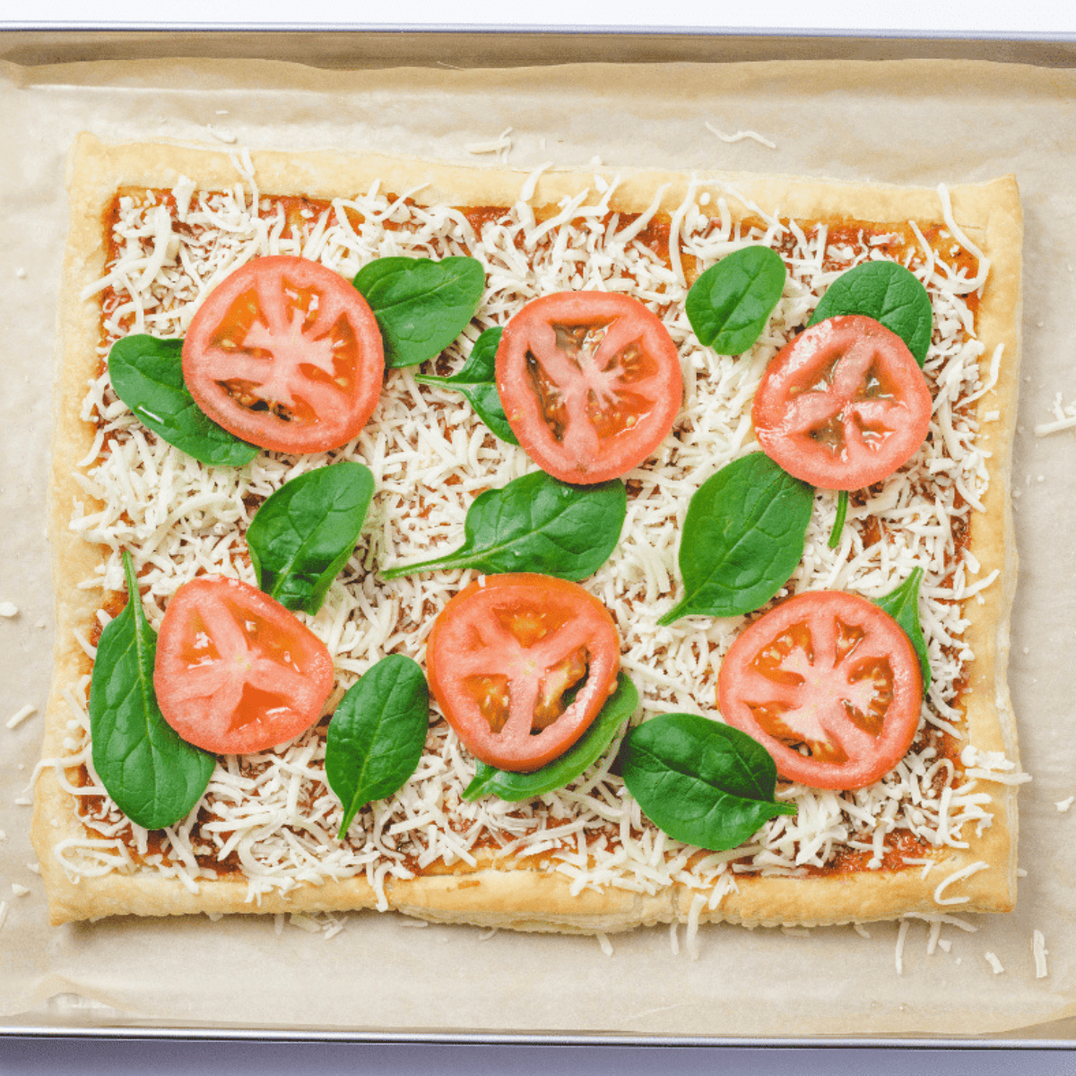 Sliced vegetables prepared in bowls next to a puffed-up pastry crust with pizza sauce being spread inside the border.