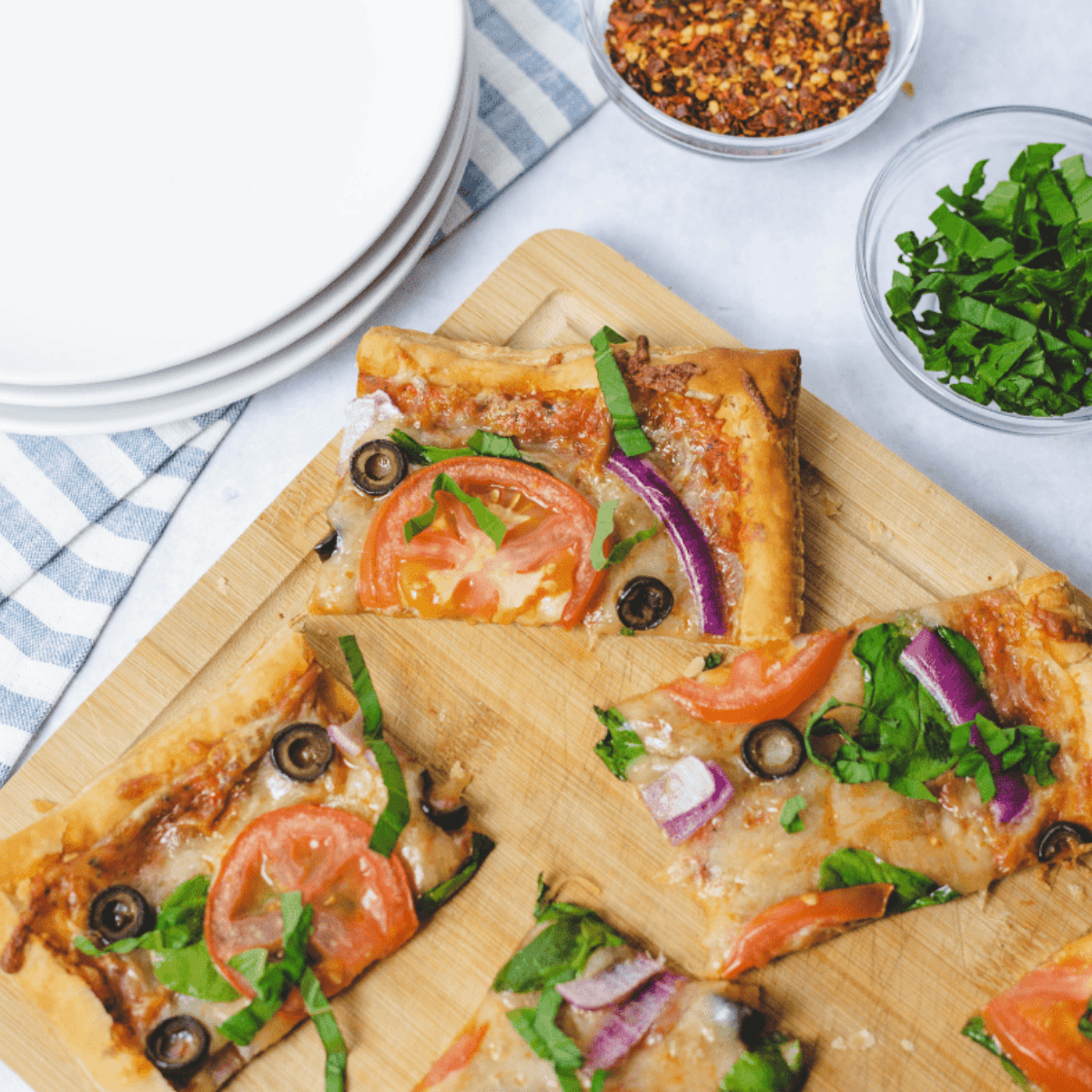 Golden puff pastry pizza topped with melted cheese, vegetables, and fresh basil, served on a cutting board.