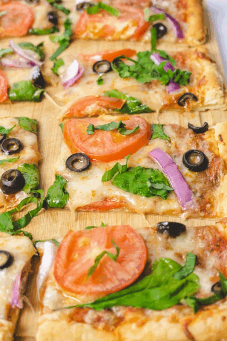 Flaky puff pastry pizza topped with melted cheese, fresh vegetables, and basil, served on a wooden board.