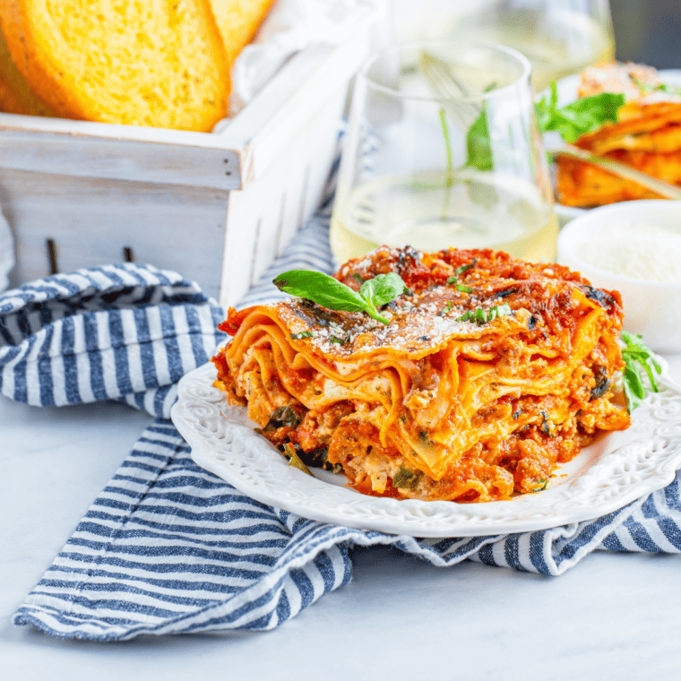 side view: a large piece of air fryer lasagna on a plate with a basket of garlic bread and a wine glass in the background