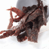 Pile of homemade air fryer beef jerky strips stacked on a rustic wooden board, glossy and dark with visible seasoning