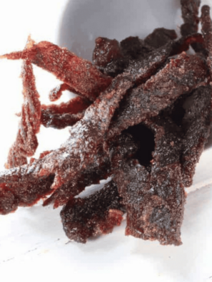 Pile of homemade air fryer beef jerky strips stacked on a rustic wooden board, glossy and dark with visible seasoning