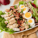 Air fryer Cobb salad with sliced chicken, bacon, hard-boiled egg, avocado, cherry tomatoes, red onion, and blue cheese on a white platter