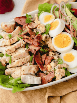 Air fryer Cobb salad with sliced chicken, bacon, hard-boiled egg, avocado, cherry tomatoes, red onion, and blue cheese on a white platter
