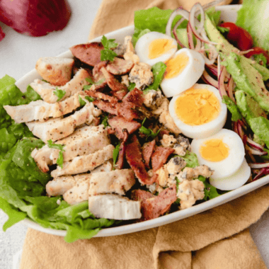 Air fryer Cobb salad with sliced chicken, bacon, hard-boiled egg, avocado, cherry tomatoes, red onion, and blue cheese on a white platter