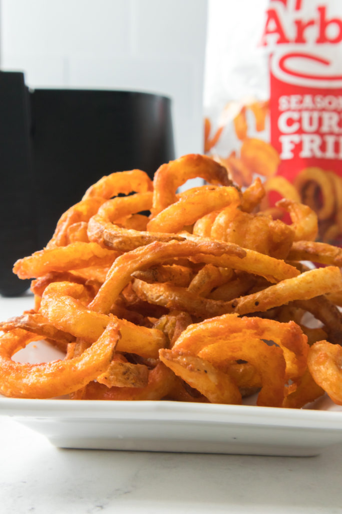 closeup of curly fries on a white plate with packaging and air fryer basket in the background