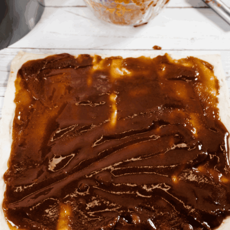 Spreading cinnamon butter mixture evenly over rolled-out puff pastry sheet.