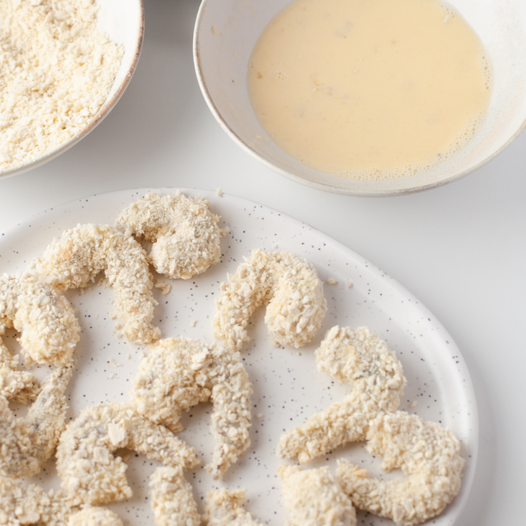 Shrimp being dipped into beaten eggs and then coated in breadcrumb mixture on a plate.