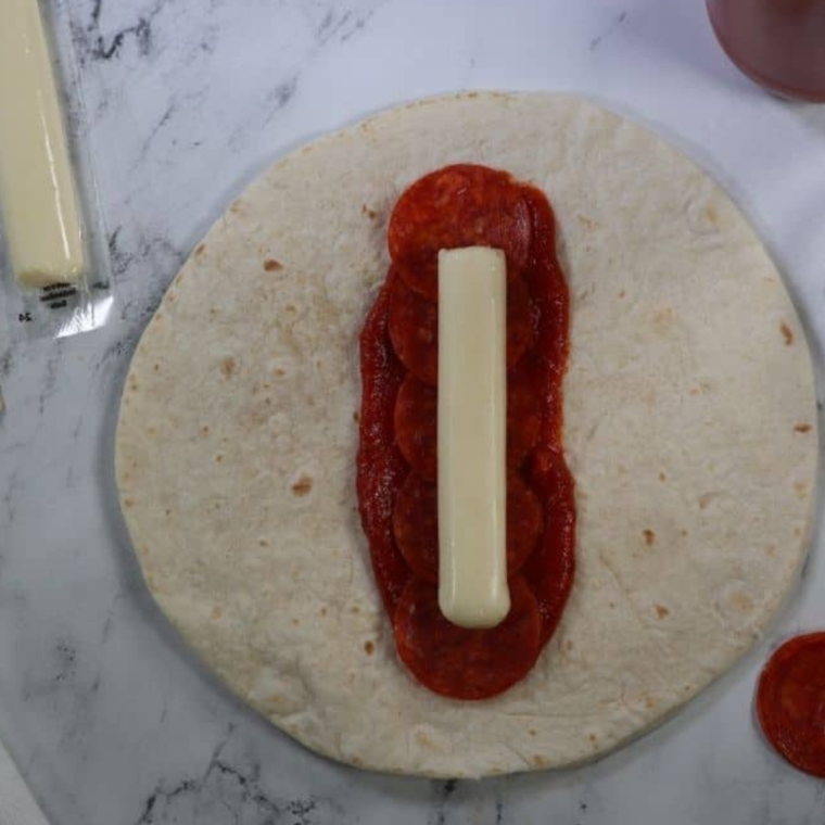 Place a smear of tomato sauce/pizza sauce in the center of the tortilla, and then add your sliced pepperoni, following by a cheesestick.