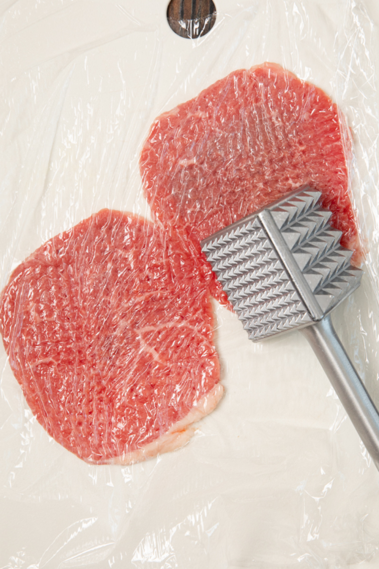 Cube steak being pounded with a meat mallet between plastic wrap to thin and tenderize before marinating.