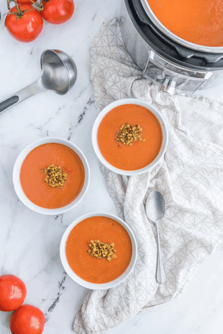 A creamy bowl of red tomato soup topped with a swirl of heavy cream and fresh basil, served in a white ceramic bowl next to the Instant Pot.
