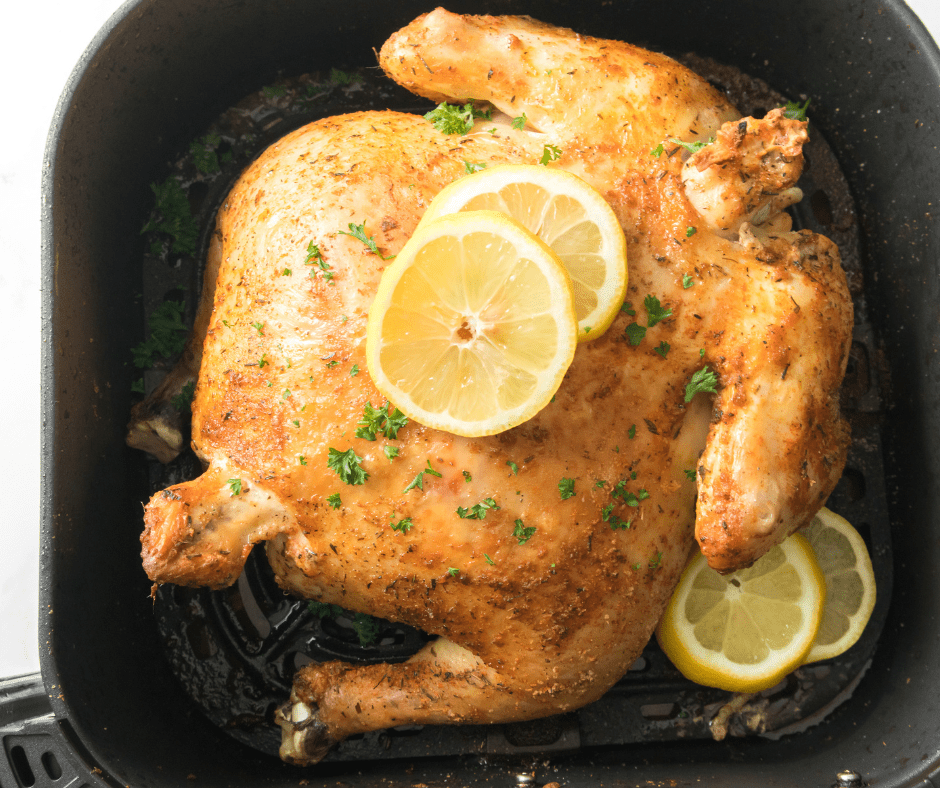 Aerial shot of cooked air fryer rotisserie chicken with two slices of lemon and herbs on top. The chicken is in a black air fryer basket and there are two lemon slices on the side.