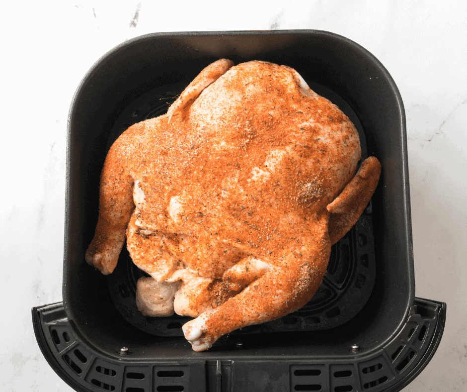 Aerial shot of a raw air fryer whole chicken rotisserie style in a black air fryer basket. There are seasonings visible on the raw chicken. 