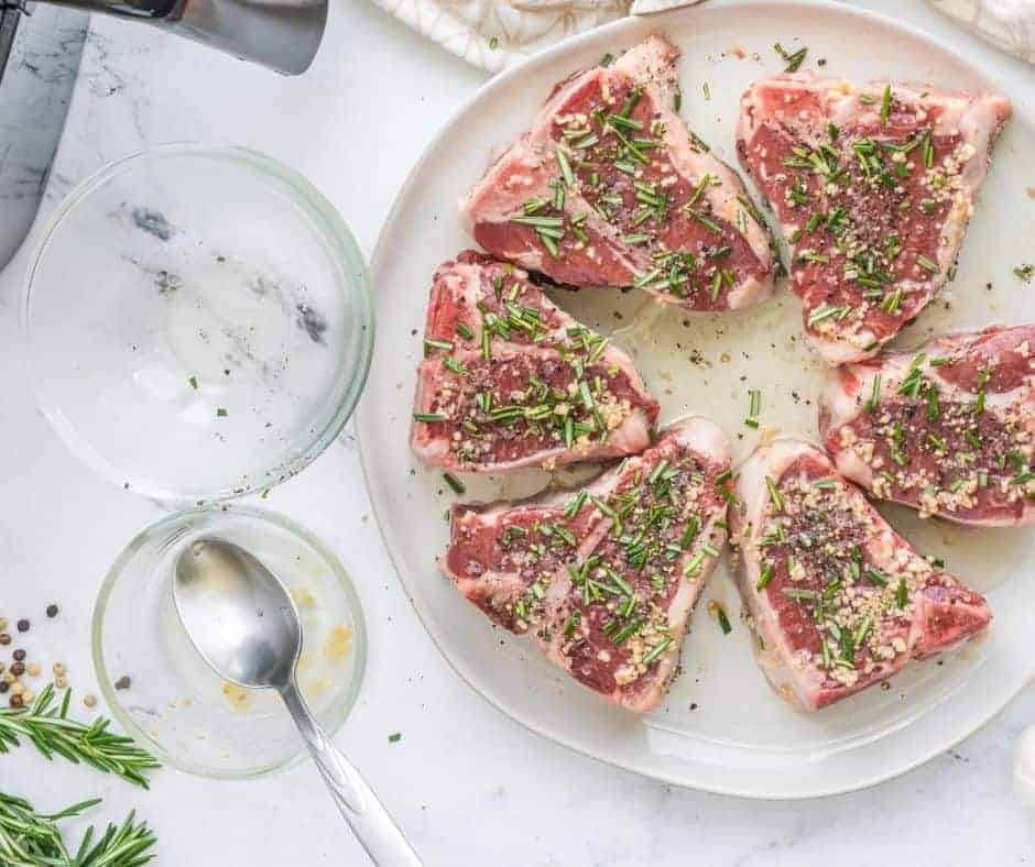An aerial view of 6 pieces of raw lamb chops on a white plate with herbs and spices on top for this easy lamb chop recipe.