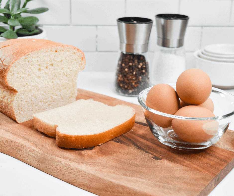 Ingredients needed for egg in bread hole. A loaf of white bread with one piece sliced off sits on a wooden cutting board with a glass bowl containing four eggs. Salt and pepper can be seen in the background.