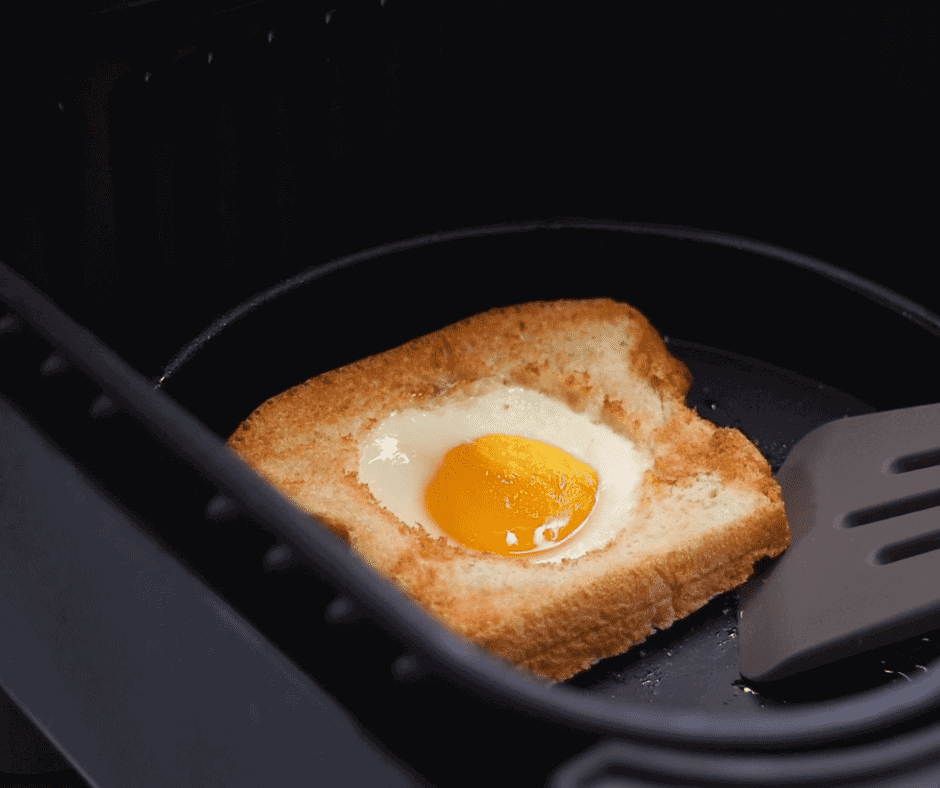 Cooked toad in the hole egg recipe in a black air fryer basket. A black spatula is lifting up one corner of the bread.