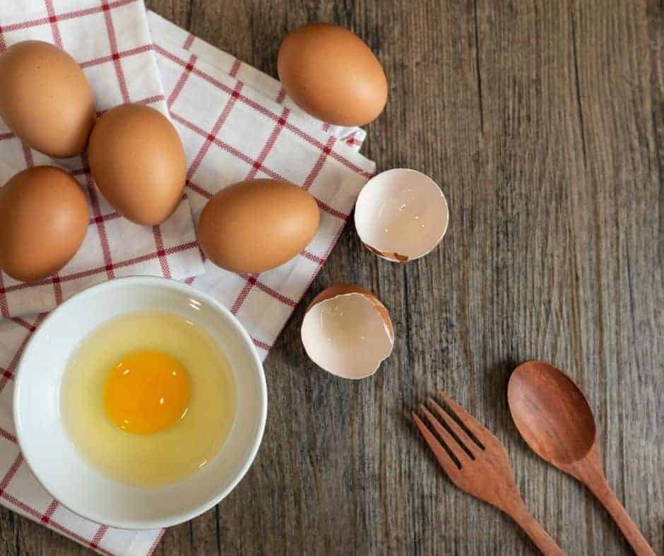 On a wooden table lay eggs on top of a kitchen towel. There is a white bowl with 1 egg inside and the shell is sitting next to the bowl on the table.