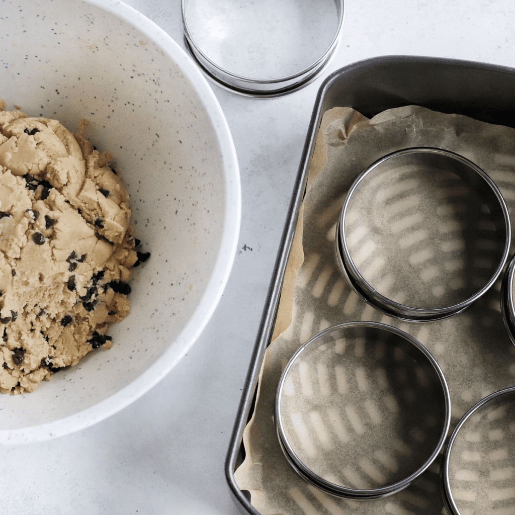 Cookie dough resting at room temperature beside an air fryer preheating to 315°F, with parchment paper lining the basket and greased muffin rings ready for use.