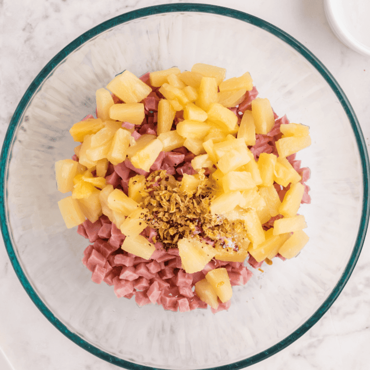Mini bagels halved on a surface with a bowl of diced ham, pineapple, ginger, red pepper flakes, salt, and pepper prepared for topping.