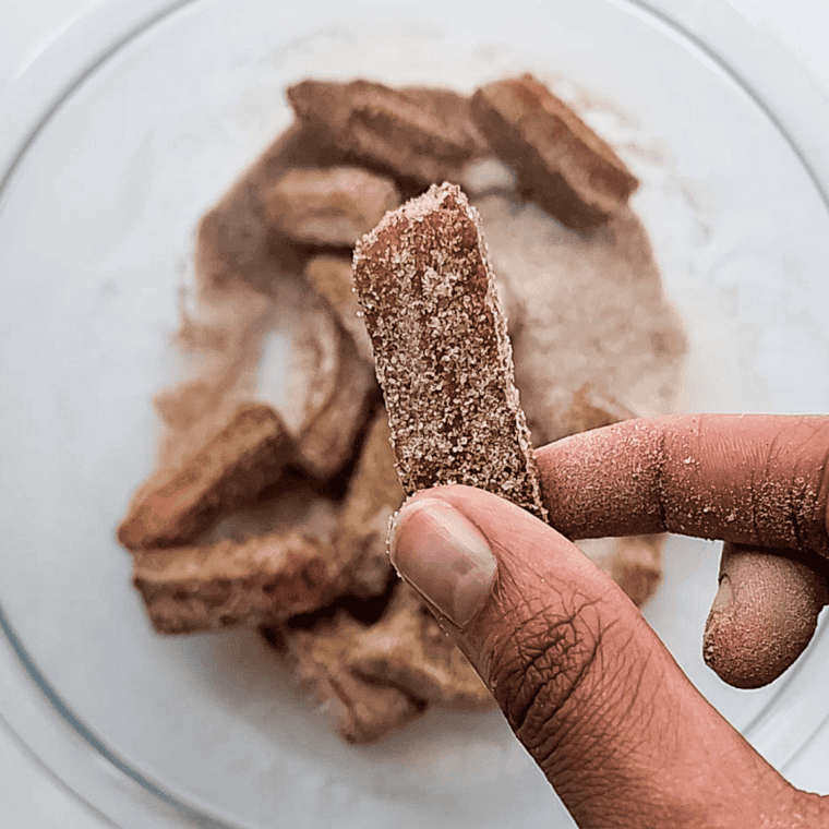 Mixing sugar and cinnamon in a bowl, coating churro bites thoroughly, and serving with a dipping sauce while warm.