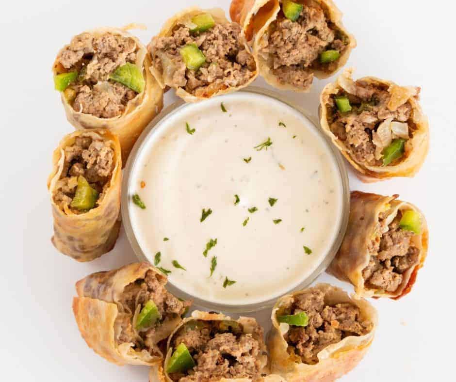 overhead: a circle of philly cheesesteak egg rolls surrounding a white dipping sauce