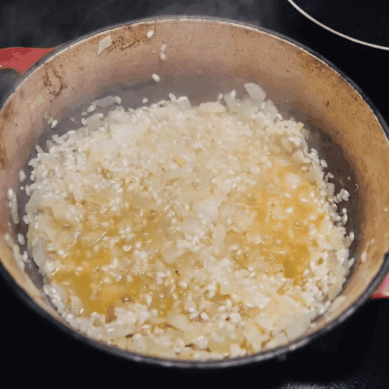 Rice cooking in a skillet with onions and wine, gradually absorbing mushroom-infused stock one ladle at a time while being stirred.