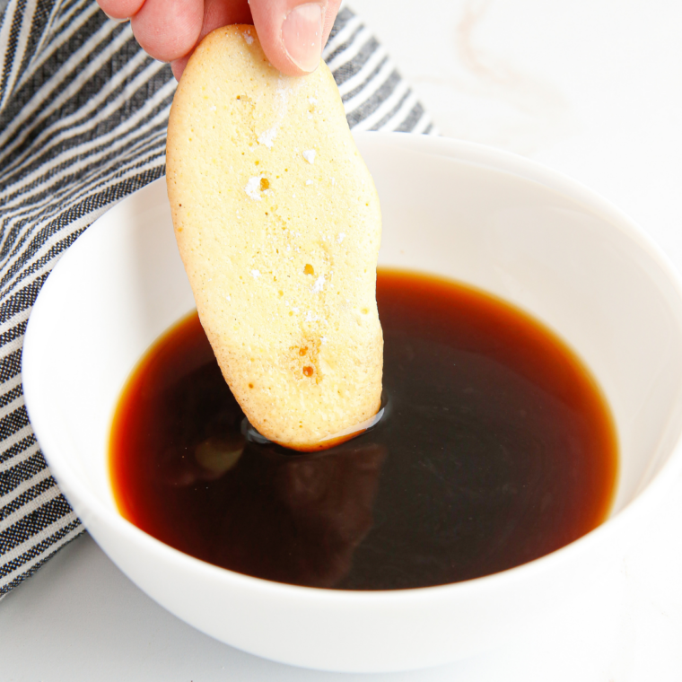 Hand dipping a ladyfinger into a shallow bowl of espresso above an 8x8 baking dish
