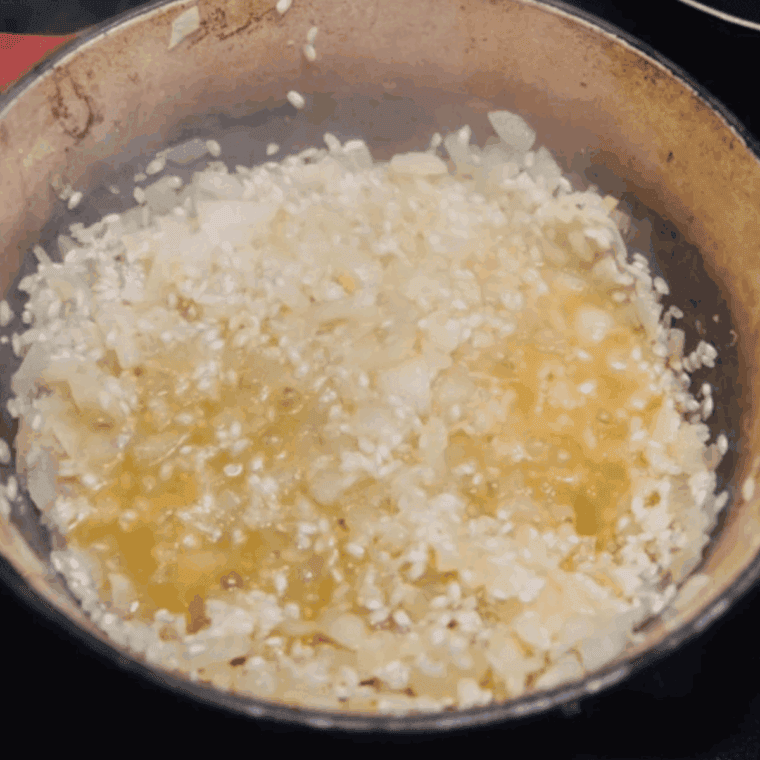 Rice cooking in a skillet with onions and wine, gradually absorbing mushroom-infused stock one ladle at a time while being stirred.