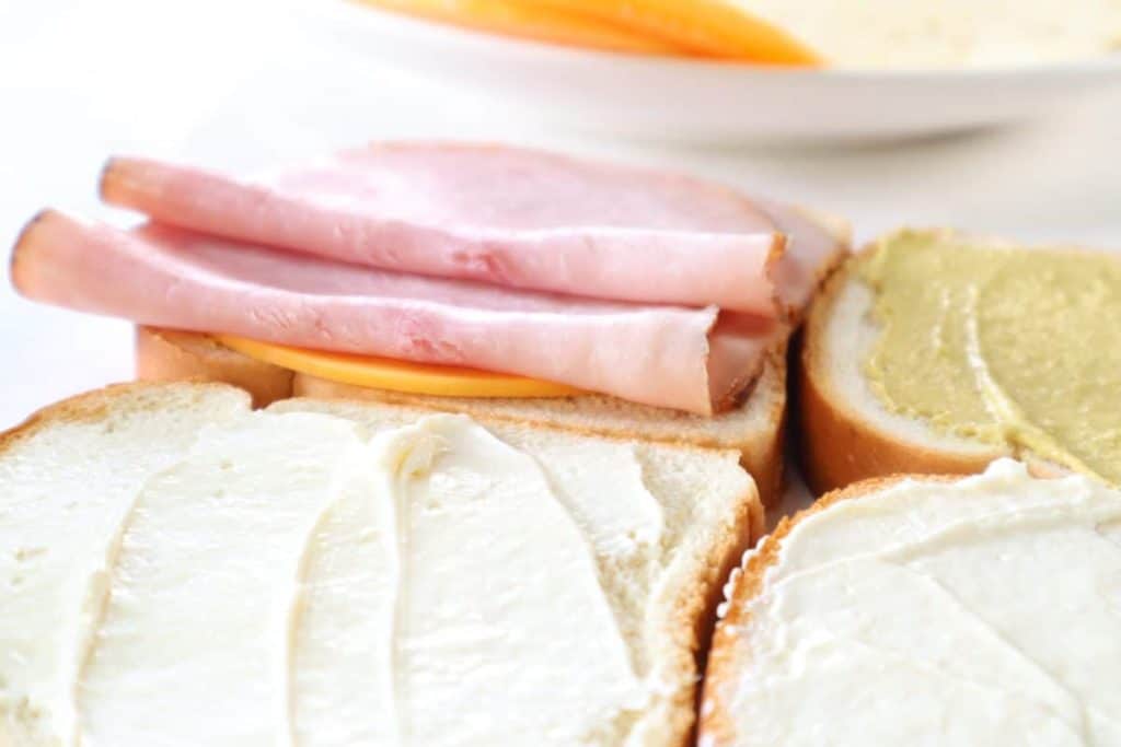 closeup: a slice of bread with mayo, and another slice with cheese and ham on it to make a grilled ham and cheese sandwich