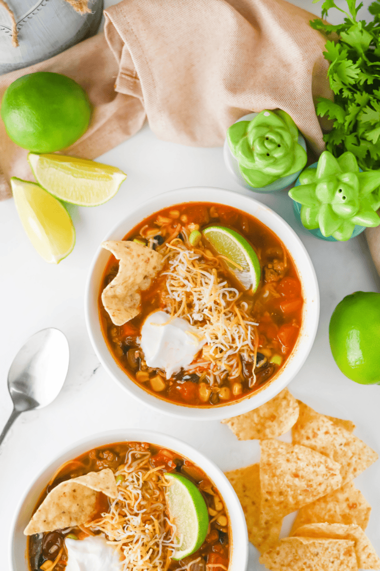 Ladling chicken enchilada soup into a bowl topped with tortilla strips, avocado, and lime.
