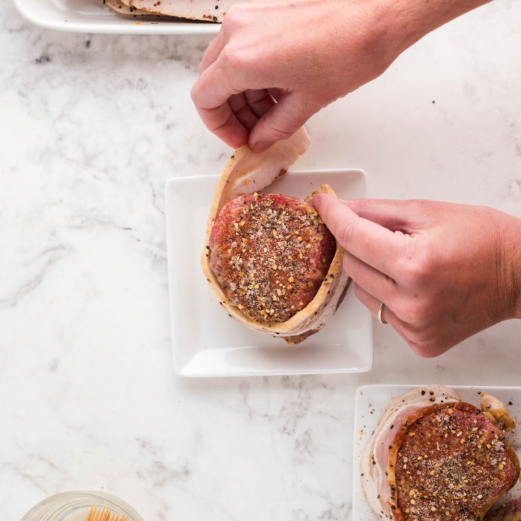 Raw steaks being patted dry with paper towels and seasoned with kosher salt and black pepper on a cutting board.