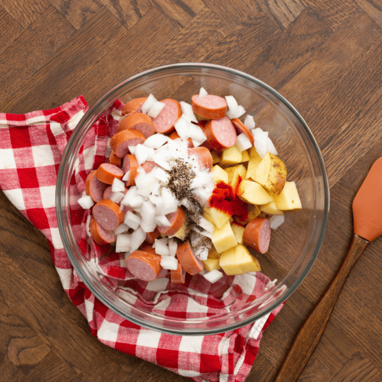 Uncooked potatoes and kielbasa mixture with spices in a mixing bowl. 