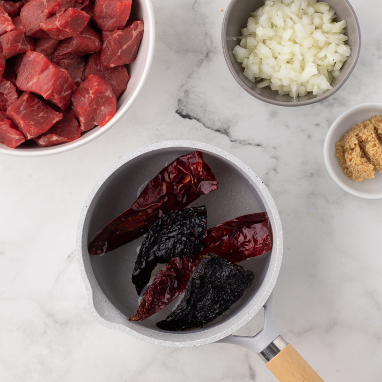 Cutting beef chuck roast into large chunks, rehydrating dried chilies, and blending them with garlic, onion, and spices for birria sauce before tossing the sauce with the beef.