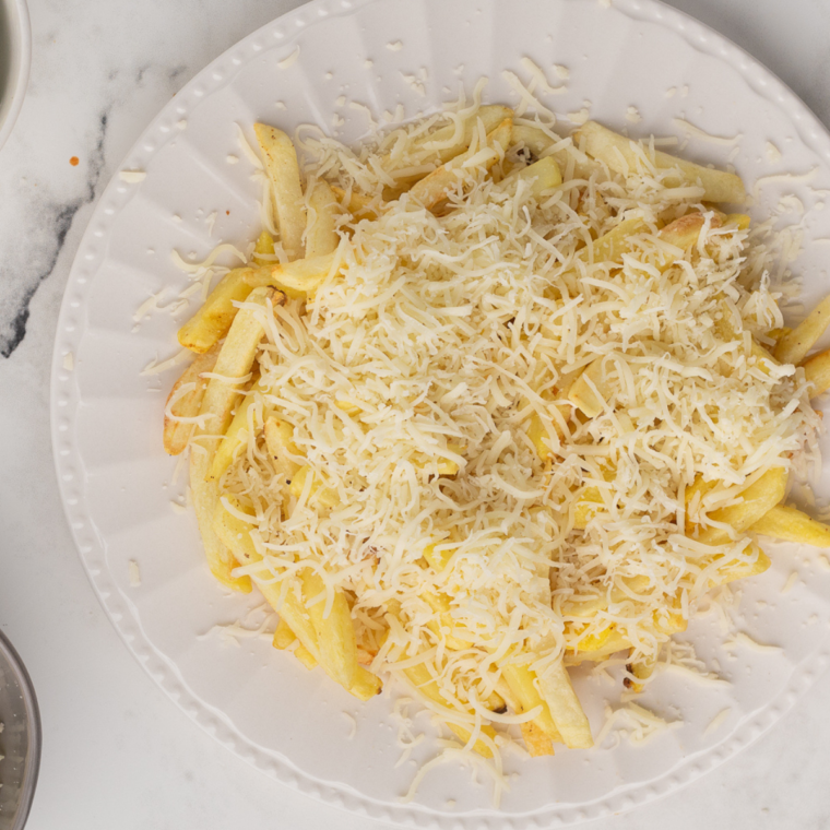 Assembling the birria fries by layering crispy fries, placing the cooked beef on top, and adding toppings like melted cheese, cilantro, onions, and a drizzle of consomé sauce.