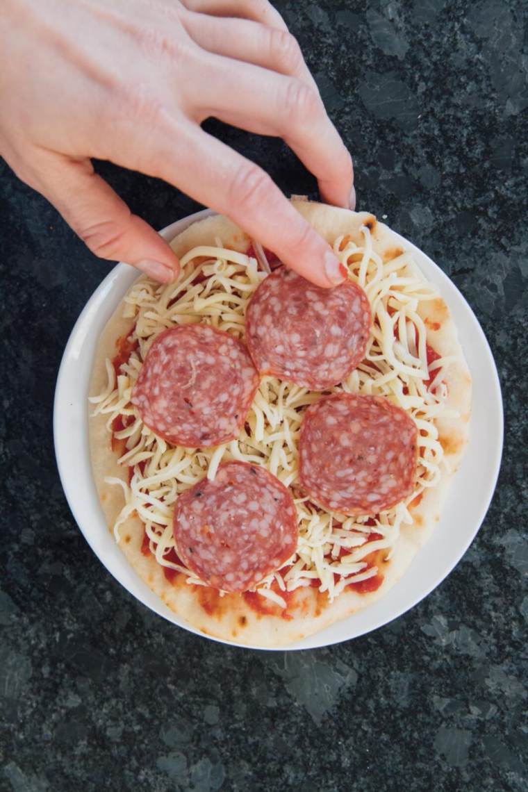 Build the pizza in the basket
Pizza sauce being spread on pre-crisped naan in the air fryer basket, then topped with shredded mozzarella cheese, pepperoni slices placed on top of the cheese, and a sprinkle of Italian seasoning and red pepper flakes